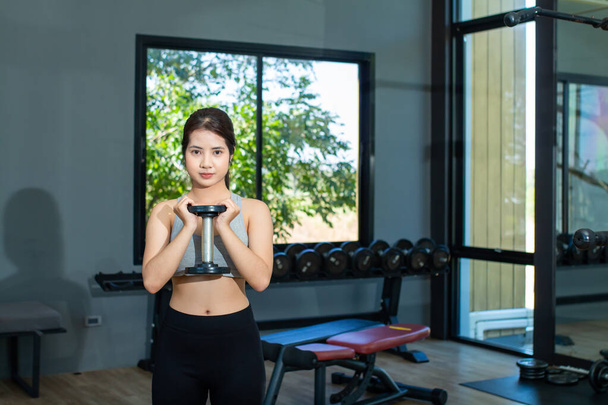 La hermosa mujer asiática está usando ropa de entrenamiento sosteniendo una mancuerna y de pie mientras mira a la cámara mientras hace ejercicios. - Foto, Imagen