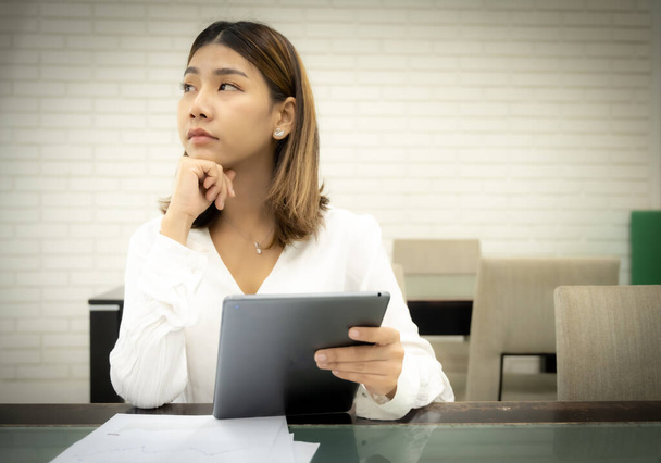 Beautiful asian business woman is sitting pretending to think about failed financial plan with a doubtful expression and holding tablet. - Foto, Bild