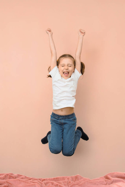 Una niña en un salto sobre una cama grita de felicidad sobre un fondo rosa. - Foto, imagen