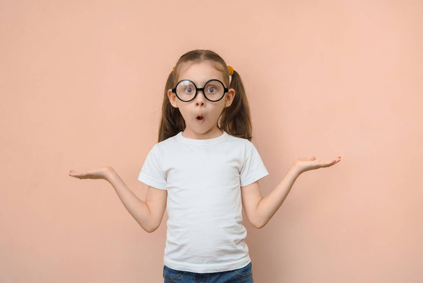 Cute funny little caucasian girl in round glasses can not choose what is in the empty space looking at the camera on a pink background. - Foto, Imagen
