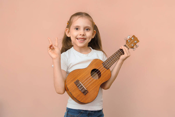 bonito caucasiano bebê menina no branco t-shirt com ukulele mostra paz gesto olha para câmera no rosa fundo. - Foto, Imagem