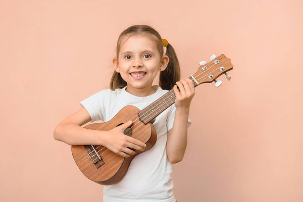 Happy funny caucasian smiling girl 6 - 8 years old in a white t-shirt with a ukulele on a pink background. - Foto, afbeelding