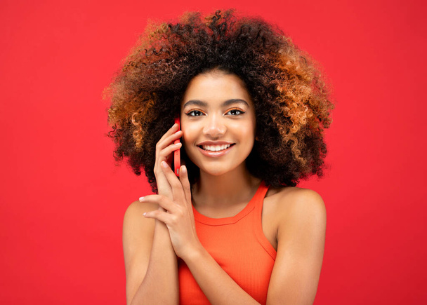 Portrait of an excited young afro american woman talking on mobile phone while standing over red color background - Fotografie, Obrázek