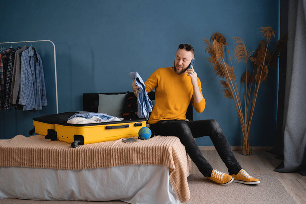 Young travel man sitting on bed near open suitcase with clothes, speaking on smartphone, booking travel tour at home, hotel reservation, buying tickets on phone. Traveler concept - Fotografie, Obrázek