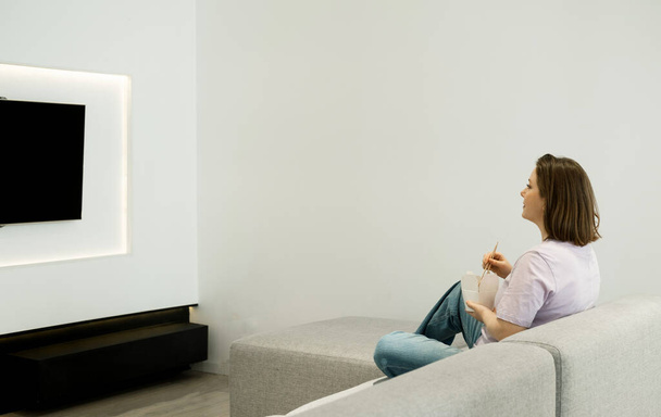 Young woman eating noodles with chopsticks, holding paper box at home in living room. Woman sitting on sofa and watching tv. - Foto, afbeelding