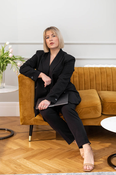 Portrait of serious gorgeous middle-aged woman businesswoman with short fair hair wearing black pantsuit, sitting on mustard yellow sofa, holding grey laptop, looking at camera. Business, technology. - Valokuva, kuva