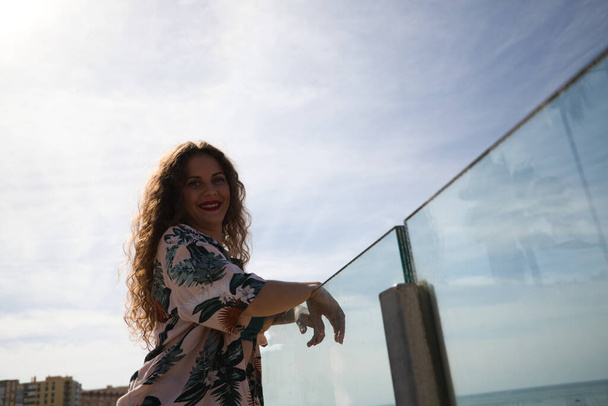 beautiful young woman, blonde with curly hair and blue eyes is leaning on the glass railing overlooking the sea. The woman has the Atlantic Ocean in front of her. In the background the blue sky. - 写真・画像