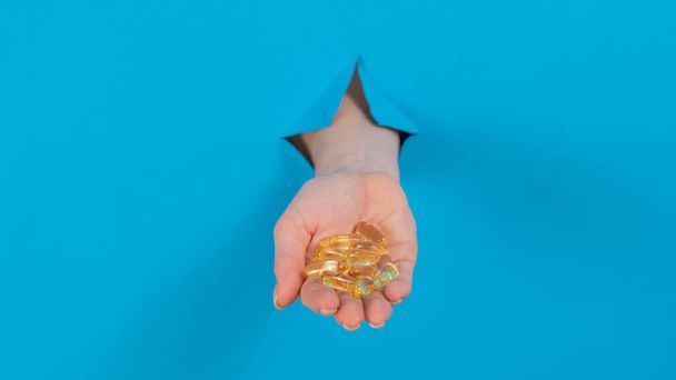 A woman holds a handful of omega 3 pills. A womans hand sticks out of a hole in a cardboard blue background - Valokuva, kuva