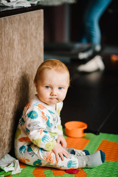 The infant is playing with toys on the rug. Funny baby boy sitting on the floor in nursery room at home. Baby health care and family concept. - Foto, afbeelding