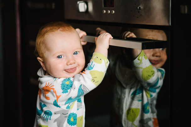 The baby learns to stand near the oven in the kitchen. Little nine months baby boy stands with support at home. Toddler just learnt how to stand on her feet. Close up. - Fotó, kép