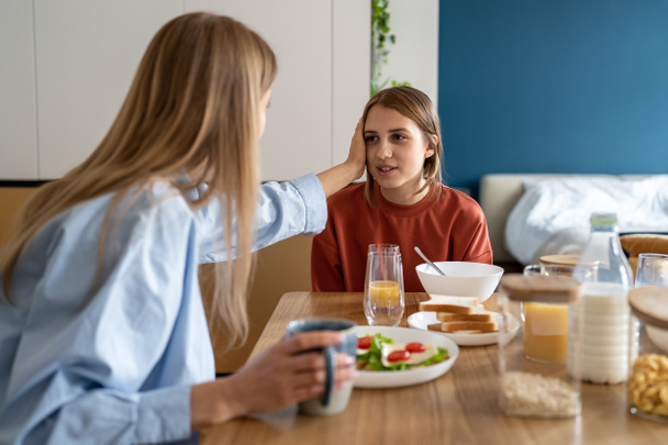 Family breakfast. Caring mother gentle touch head daughter talk about day plan. Cute teenage girl sharing with loving mom secrets while morning routine sitting at kitchen table. Trusting relationship - Photo, Image