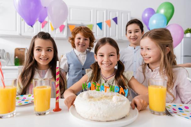 niños felices mirando a la cámara cerca de pastel de cumpleaños y globos durante la celebración en casa  - Foto, Imagen