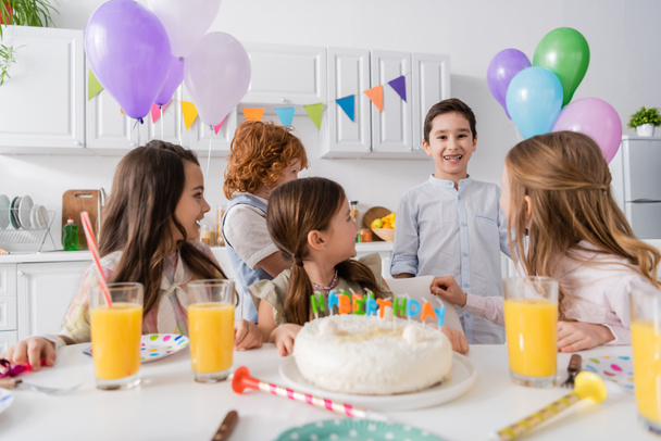 wesoły dzieci patrząc na szczęśliwy solenizant z aparatem na zębach w pobliżu tortu ze świecami na stole  - Zdjęcie, obraz