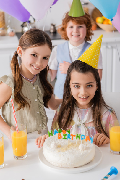 パーティーキャップの幸せな女の子誕生日ケーキの近くに友人とカメラを見て  - 写真・画像
