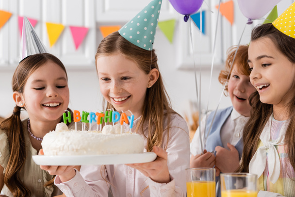 felice ragazza che tiene torta di compleanno con candele vicino ragazze allegre durante la celebrazione a casa  - Foto, immagini