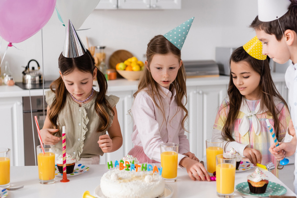 preteen κορίτσια σε καπελάκια κόμμα κοιτάζοντας τούρτα γενεθλίων δίπλα στο χυμό πορτοκάλι στο τραπέζι  - Φωτογραφία, εικόνα