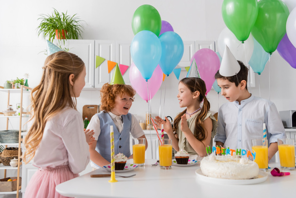 filles heureuses dans les casquettes de fête applaudissant les mains et chantant chanson joyeux anniversaire près des garçons et gâteau  - Photo, image