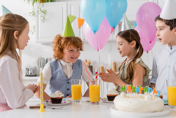 Vorschulmädchen in Partymützen klatschen in die Hände und singen neben Jungen und Kuchen ein Happy Birthday-Lied  - Foto, Bild
