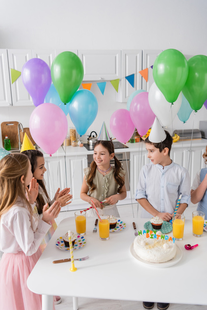 χαρούμενα παιδιά χειροκροτούν και τραγουδούν χαρούμενο τραγούδι γενεθλίων δίπλα στην τούρτα με κεριά και μπαλόνια  - Φωτογραφία, εικόνα