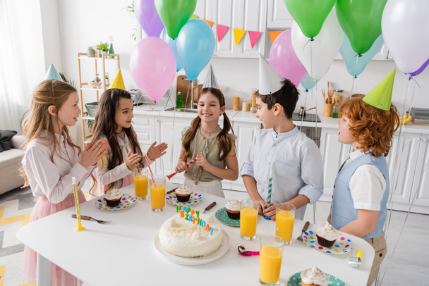 positieve kinderen klappen handen en zingen gelukkige verjaardag lied naast taart met kaarsen en ballonnen  - Foto, afbeelding