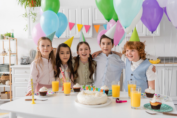 Positive Kindergruppe singt Happy Birthday Song neben Kuchen mit Kerzen und Luftballons  - Foto, Bild