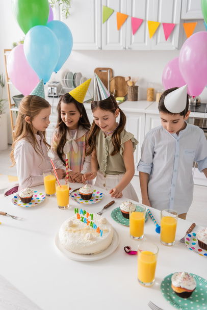 happy preteen girls in party caps smiling next to boy and cupcakes during birthday party  - Fotoğraf, Görsel