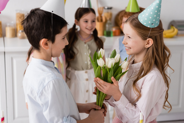menino pré-adolescente positivo dando tulipas para menina de aniversário satisfeito perto de amigos no fundo borrado  - Foto, Imagem