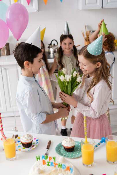 preteen boy giving tulips to cheerful birthday girl near cupcakes and friends on blurred background  - Photo, Image