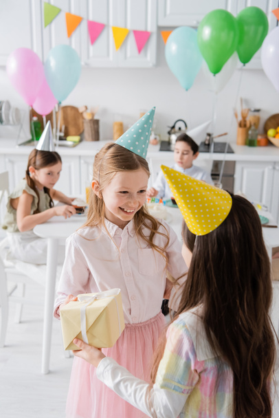 positive birthday girl in party cap receiving present from friend  - Foto, Imagem