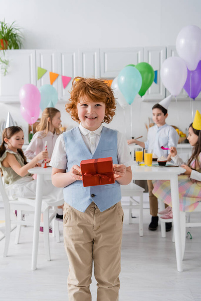 happy redhead boy holding birthday present near friends during party at home  - Foto, Imagen