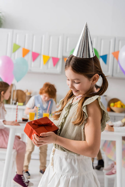gelukkig meisje in party cap holding verjaardag cadeau in de buurt van vrienden op wazig achtergrond  - Foto, afbeelding