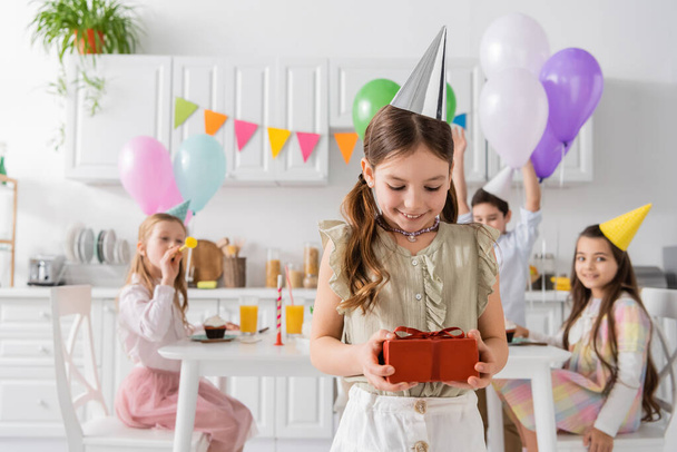 zadowolona dziewczyna w partii czapka gospodarstwa prezent urodzinowy w pobliżu przyjaciół na zamazanym tle  - Zdjęcie, obraz