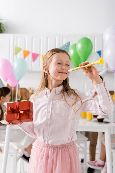 happy preteen girl with closed eyes holding birthday present and party horn near friends on blurred background  - Photo, Image