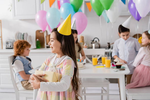 cheerful birthday girl in party cap holding gift box near friends on blurred background - Photo, Image