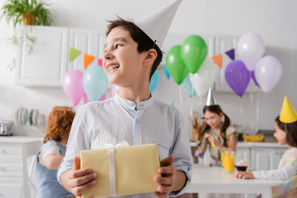χαρούμενο έφηβος αγόρι σε σιδεράκια κρατώντας δώρο γενεθλίων κοντά σε φίλους σε θολή φόντο  - Φωτογραφία, εικόνα