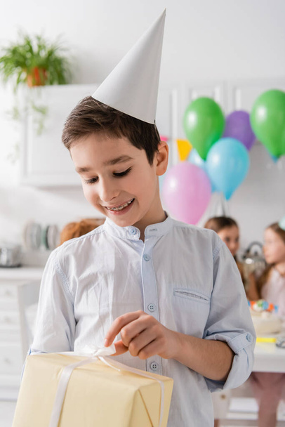 sorridente preteen ragazzo in bretelle tenere regalo di compleanno vicino amici su sfondo sfocato  - Foto, immagini