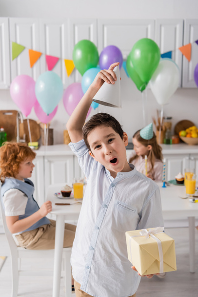 funny preteen boy in braces holding birthday present and adjusting party cap near friends on blurred background  - Foto, afbeelding