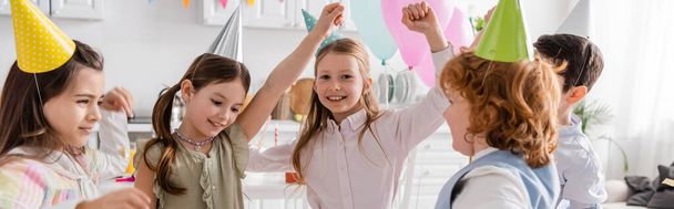 cheerful children in party caps dancing during happy birthday celebration at home, banner  - Foto, Imagen