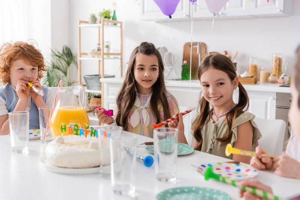 boldog lányok és fiú gazdaság party szarvak mellett születésnapi torta és nézi barát elmosódott előtérben  - Fotó, kép