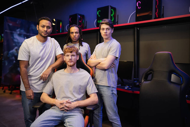 Multiethnische Spieler in grauen T-Shirts blicken in Cyber-Club in die Kamera  - Foto, Bild
