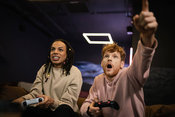 KYIV, UKRAINE - FEBRUARY 13, 2023: Excited man pointing with finger while playing video game with multiracial friend in gaming club  - Foto, afbeelding