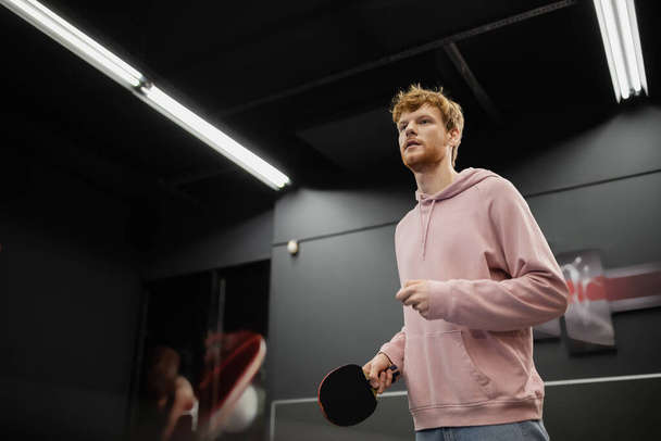 Young redhead man with racket playing table tennis in gaming club  - Fotó, kép
