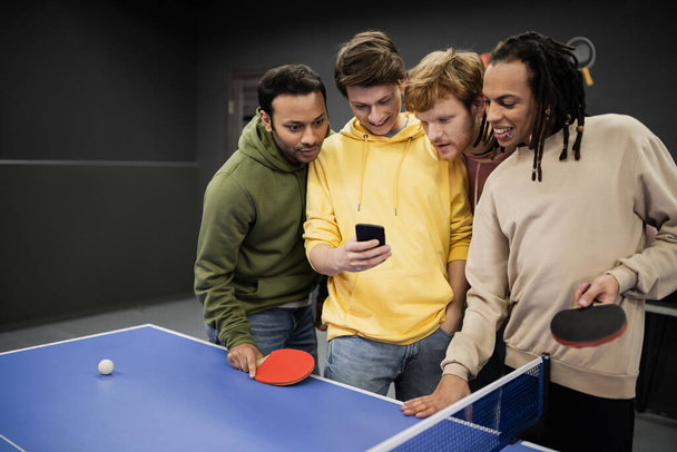 Amigos interracial sonrientes usando teléfono inteligente cerca de tenis de mesa en el club de juegos  - Foto, imagen