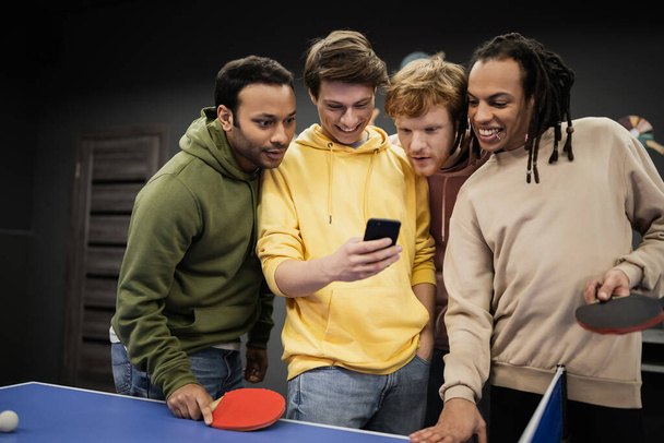Wieloetniczni przyjaciele uśmiechnięci podczas korzystania ze smartfona w pobliżu tenisa stołowego w klubie gier  - Zdjęcie, obraz