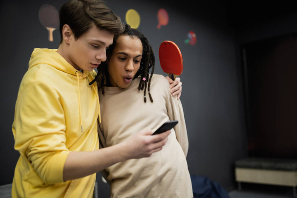 Молодий чоловік використовує смартфон і тримає тенісну ракетку біля шокованого багаторасового друга в ігровому клубі
  - Фото, зображення