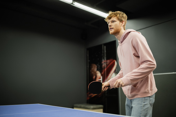 Young redhead man holding racket while playing table tennis in gaming club  - Fotografie, Obrázek