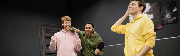 Cheerful interracial men with tennis racket showing yes gesture near friend in gaming club, banner  - Photo, Image