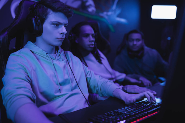 Zaměřený muž ve sluchátkách hraní videohry v blízkosti rozmazané multietnické přátele v kybernetickém klubu  - Fotografie, Obrázek