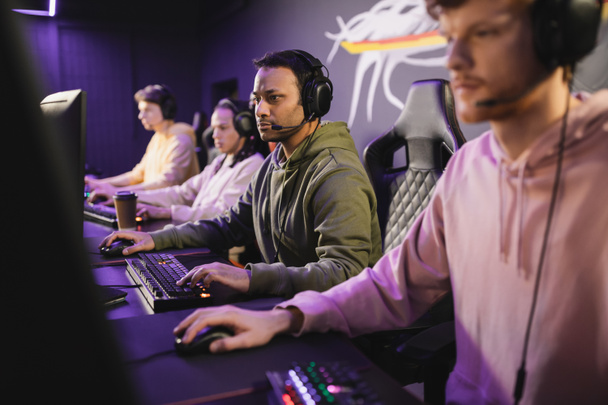Indian gamer in headphones playing video game on computer near blurred team in cyber club  - Foto, Bild