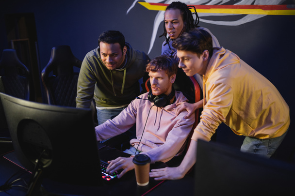 Multiethnic friends playing video game near coffee to go in gaming club  - Foto, imagen